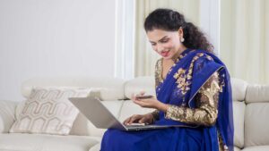 An Indian woman in a traditional blue and gold sari sits on a white couch, smiling as she looks at her laptop while holding a credit card, seemingly making an online purchase.