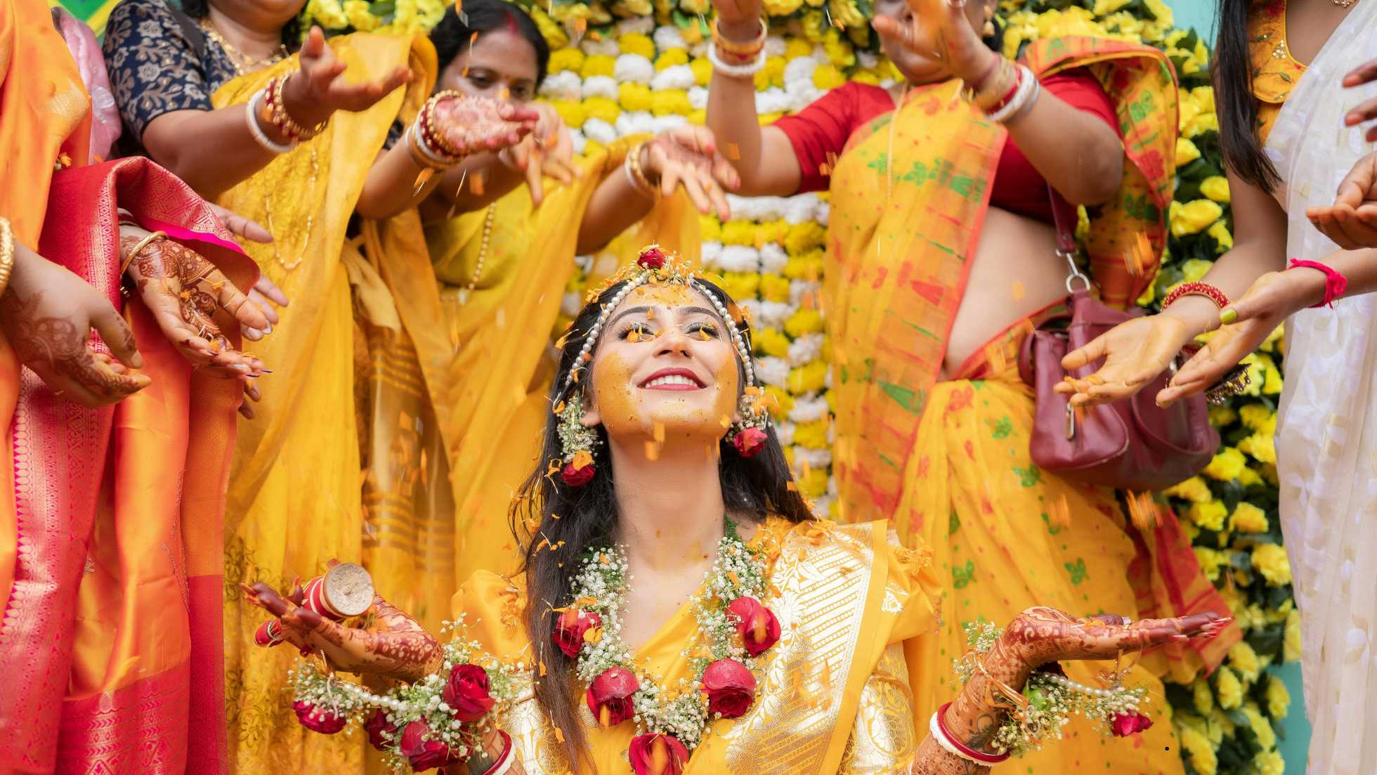 Joyful Indian bride during a Haldi ceremony, smiling with turmeric paste on her face and red and white flower garlands, as women shower her with yellow powder.