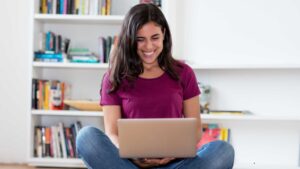 A happy young woman sitting cross-legged on the floor, smiling as she uses her laptop to read a review of the Flipkart SBI Credit Card and its benefits.