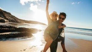 Happy couple enjoying a piggyback ride on a sunny beach at sunset, laughing with arms raised, celebrating vacation
