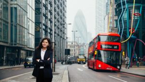 A confident NRI woman standing in a busy London financial district with modern buildings and a red double-decker bus, representing global Indians exploring NRI credit cards in India for travel, international spending, and financial convenience.